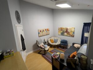 Current photo of the Corner Office. Point of view from desk area looking towards half the office set up as a sitting area, with a couch and 2 types of chairs